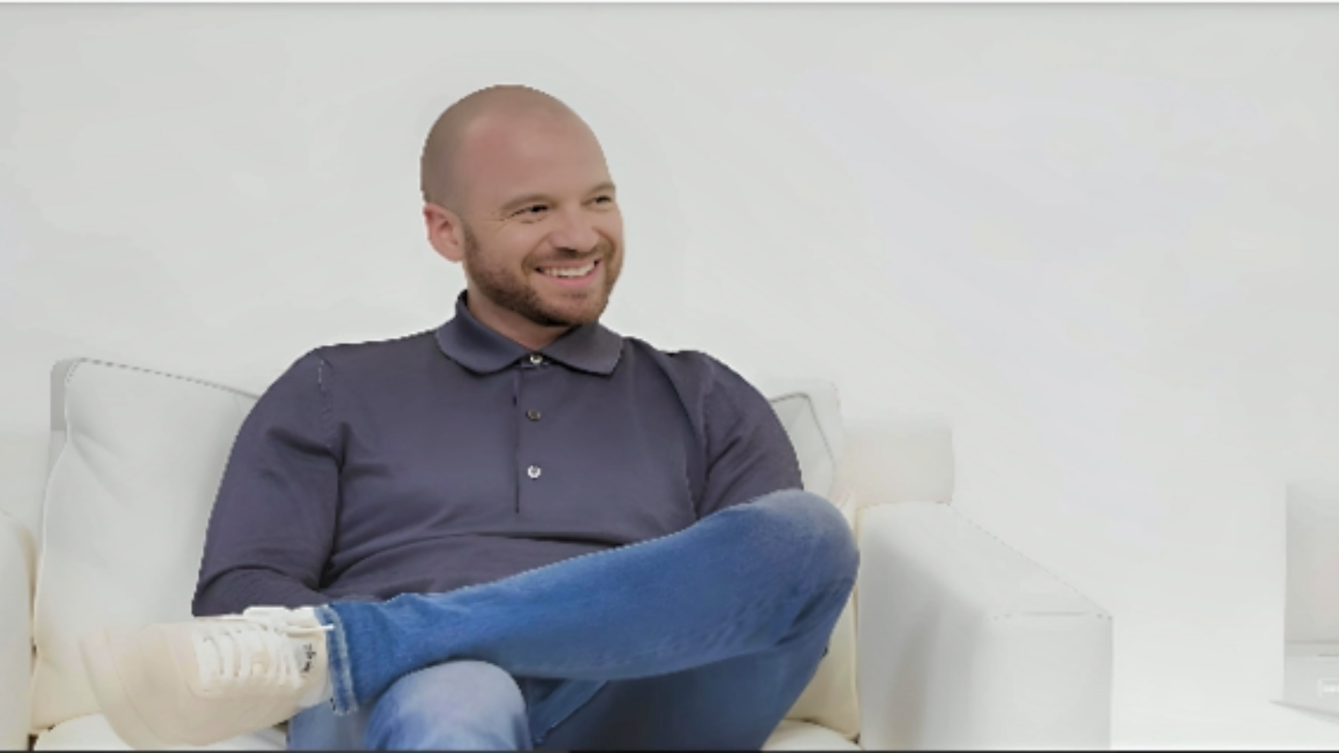 Sean Evans sitting on sofa wearing white sneakers, blue jeans and grey shirt. Mesmerizing smile on his face, depicting Sean Evans net worth: career and earnings