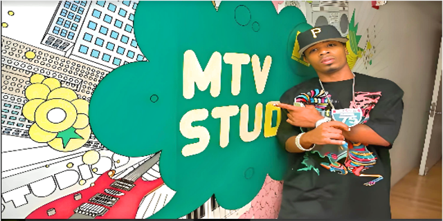 Algernod Lanier Washington aka Plies, standing in front of MTV Studio poster and posing in black shirt, depicting his successful professional career as rapper