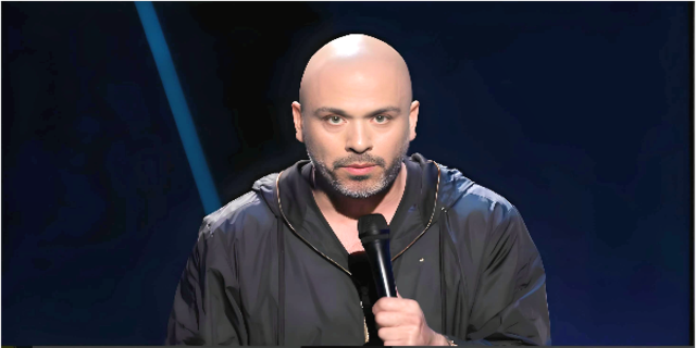 Mesmerizing picture of Jo Koy wearingblack shirt and black hoodie holding mic. 