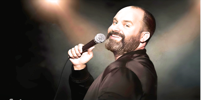 Smiling photograph of Tom Segura holding mic and wearing black coat, depicting his successas comedian.